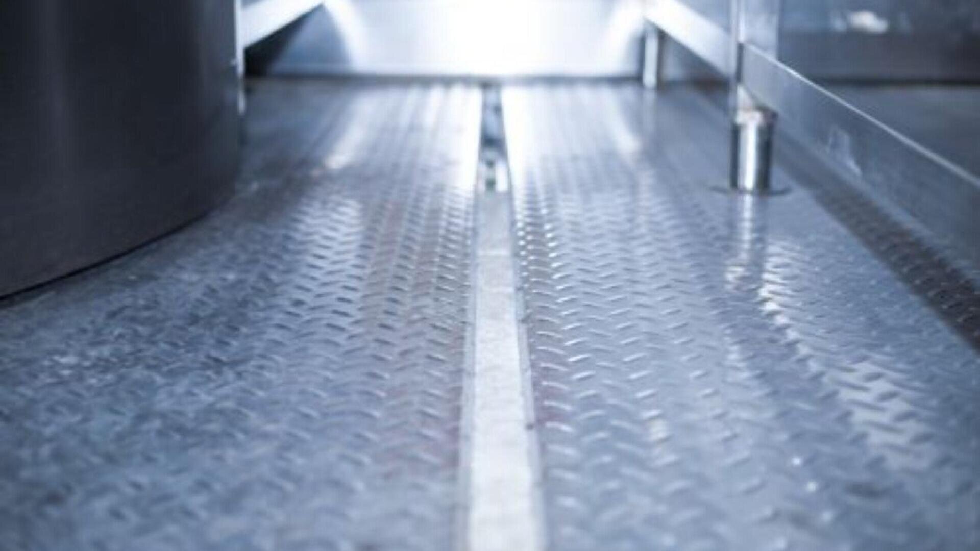 Nahaufnahme eines metallischen, rautenförmigen Bodens zwischen zwei Industriemaschinen oder -strukturen, mit reflektierenden Oberflächen und einer in Längsrichtung verlaufenden Mittelnaht.