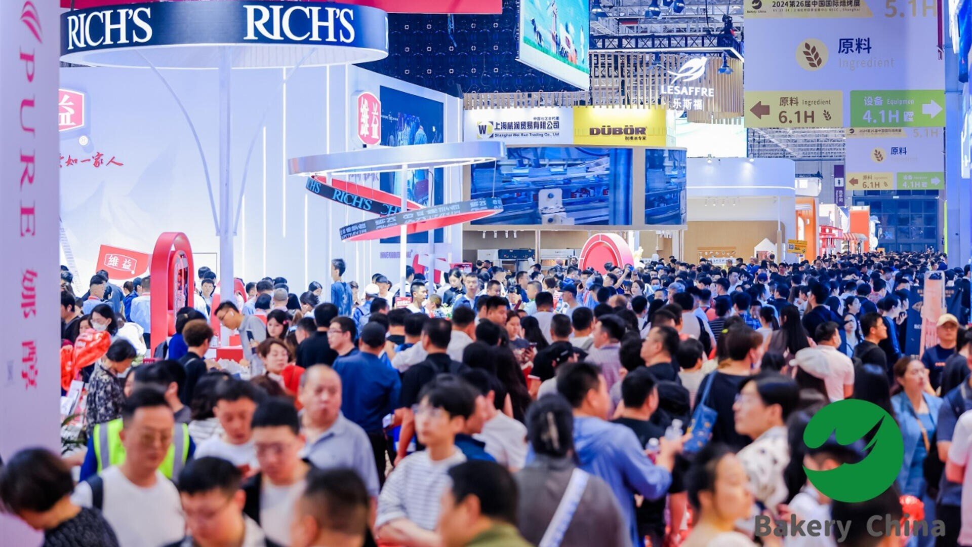 A large crowd of people fills a brightly lit exhibition hall with company booths and banners, including “Rich’s,” at a busy trade show or convention. The event is lively and bustling with attendees.