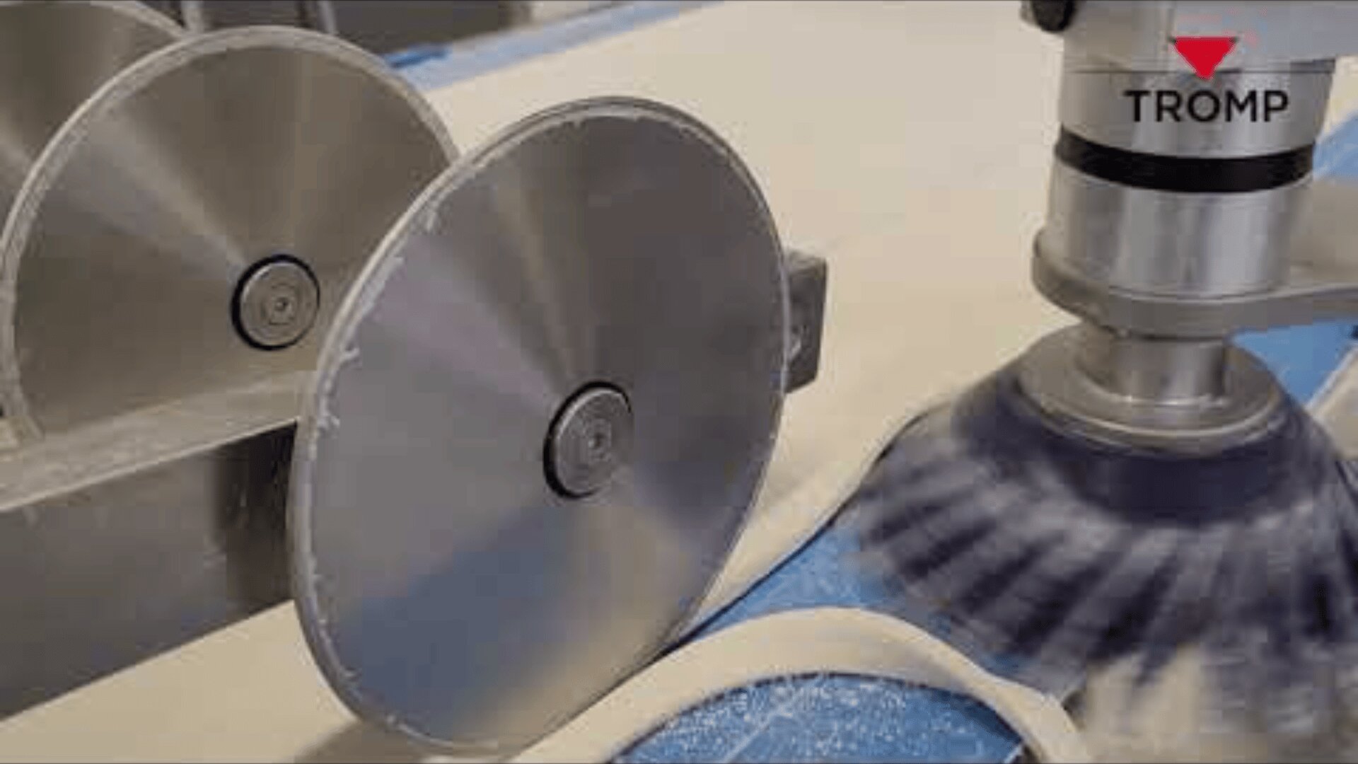 Close-up of an industrial dough-cutting machine with metal circular blades slicing dough strips on a conveyor belt. The logo TROMP appears in the top right corner.