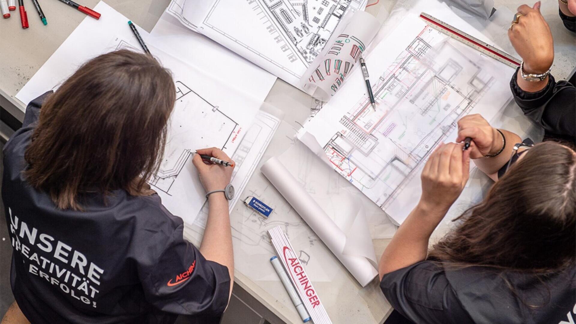 Two people in dark shirts sit at a table working with architectural blueprints, sketches, and drawing tools. Their focus is on detailed floor plans, and various stationery items are scattered around.