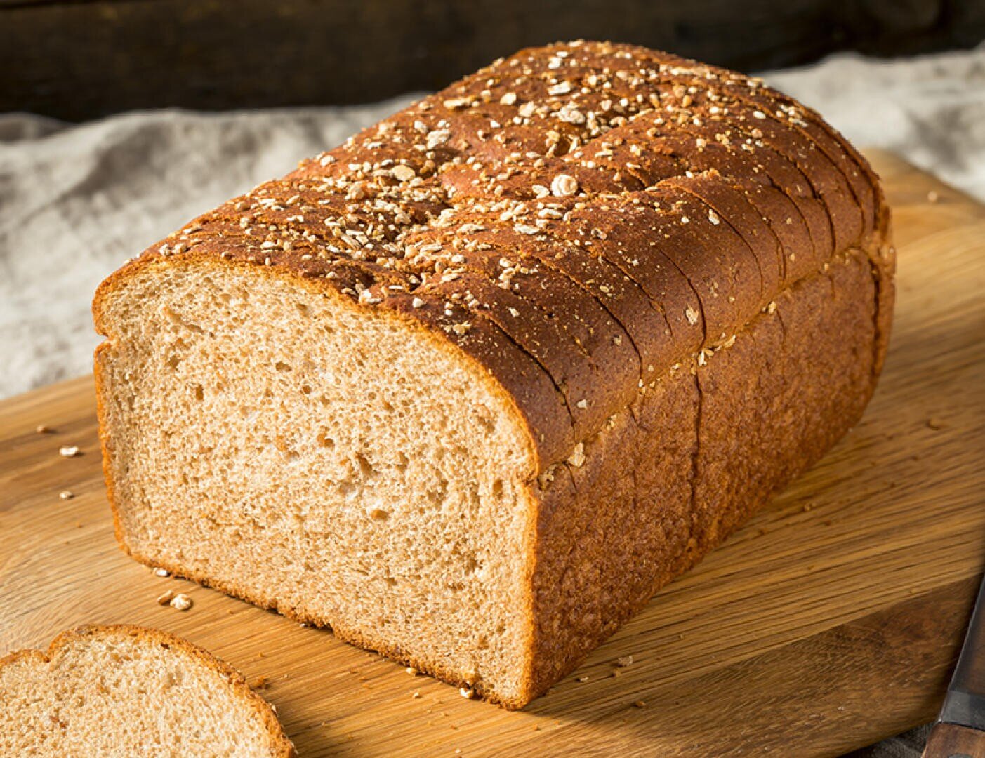 On a wooden cutting board lies a slice of wholemeal bread with oat flakes, one slice of which is partially cut off.