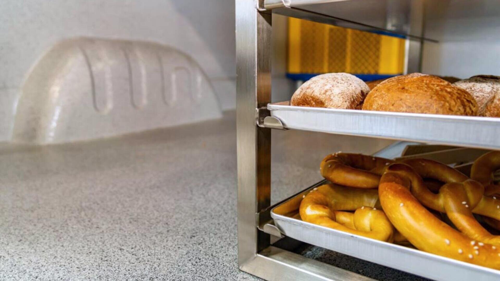 Metal trays filled with pretzels and bread rolls are stacked on a rack in a clean kitchen or bakery setting, with a loaf-shaped imprint visible on the wall in the background.