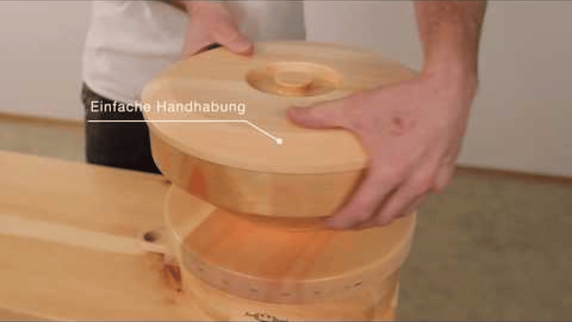 Eine Person hebt den hölzernen Deckel von einem runden Behälter ab. Auf einem Etikett in deutscher Sprache steht "Einfache Handhabung". Der Behälter und der Deckel haben eine glatte, natürliche Holzoberfläche.