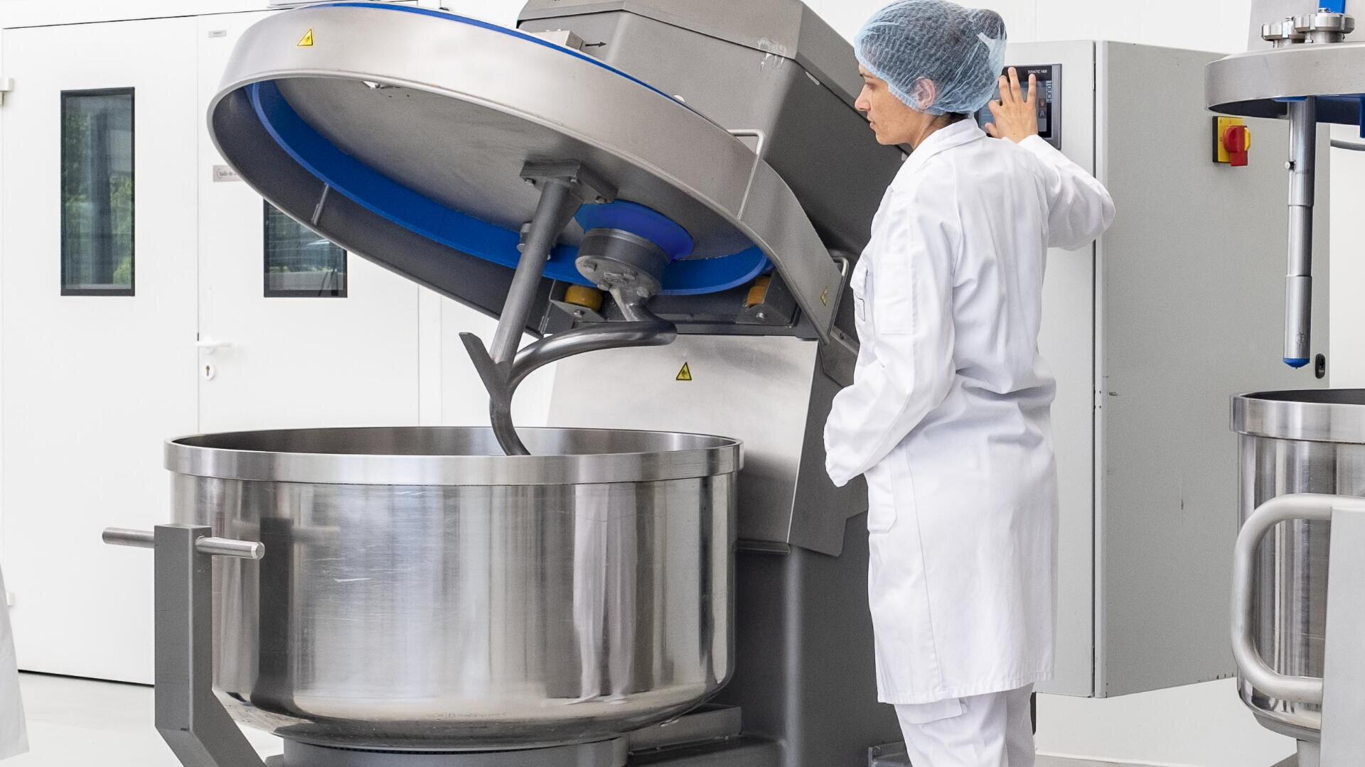 A person in a white lab coat and hairnet operates a large industrial mixing machine in a clean, modern food processing facility.