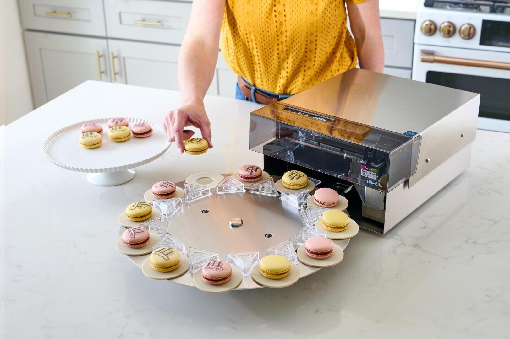 A person places a macaron on a rotating tray connected to a machine that prints motifs on the macarons. More decorated and plain macarons can be seen on a cake stand and the tray in a modern kitchen.