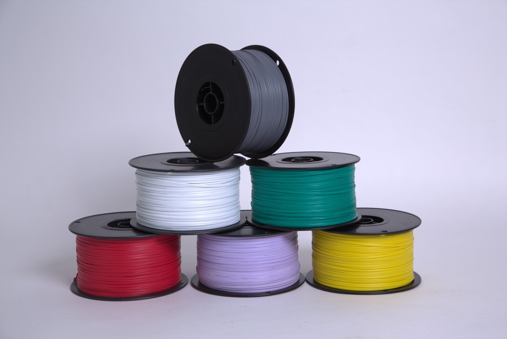Sechs Spulen mit 3D-Druckerfilament in Rot, Weiß, Grau, Grün, Lila und Gelb sind pyramidenförmig auf einem schlichten weißen Hintergrund gestapelt.