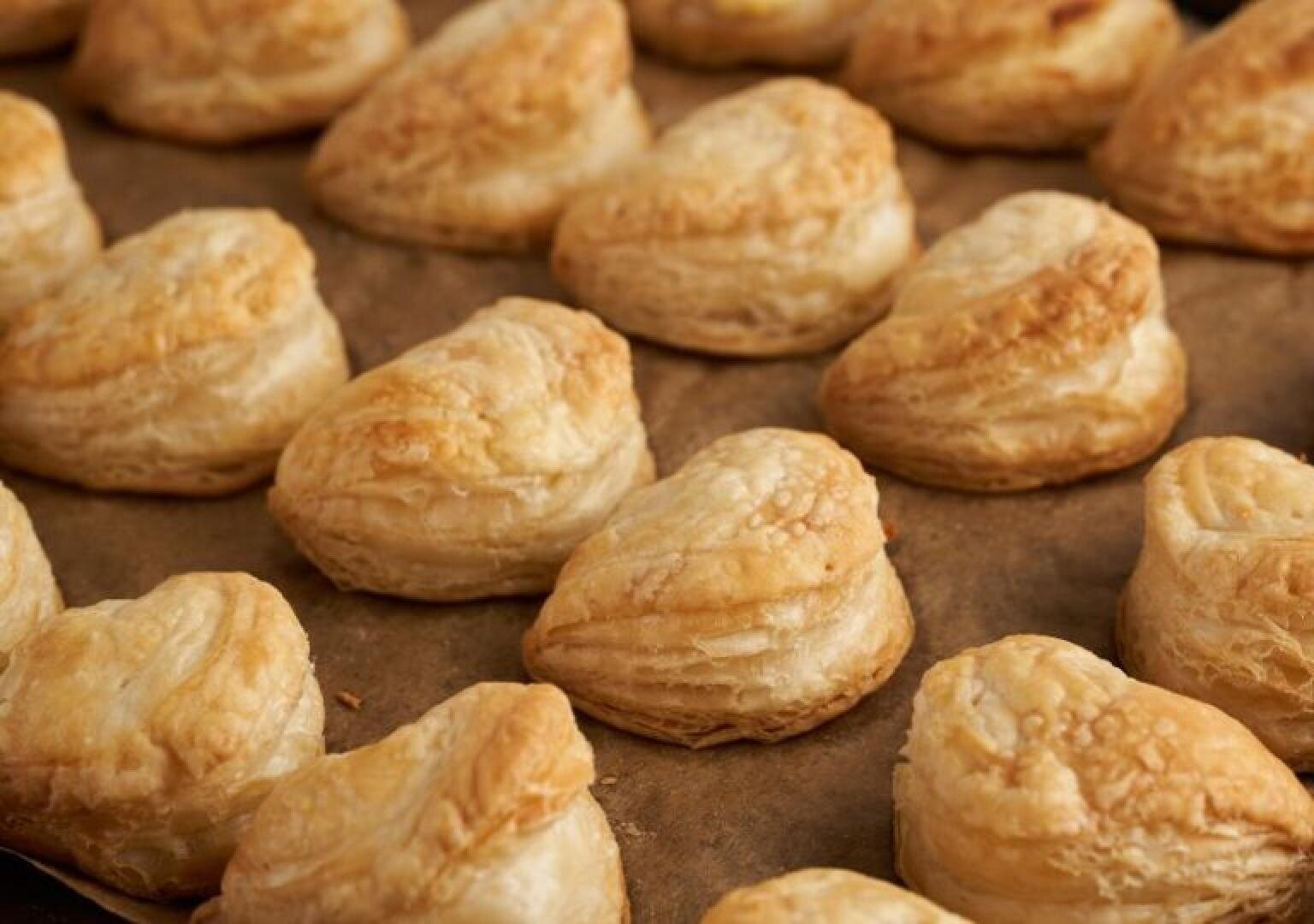 Rows of golden, heart-shaped puff pastries are arranged on a sheet of brown parchment paper, showing flaky, layered textures and a light, crispy surface.