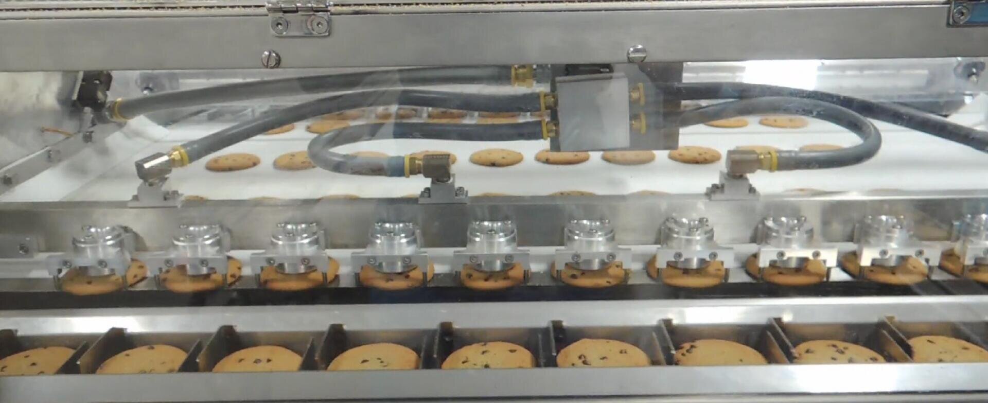 A close-up view of a cookie production line, showing chocolate chip cookies being processed by an automated machine with metal parts, tubes, and conveyor belts.