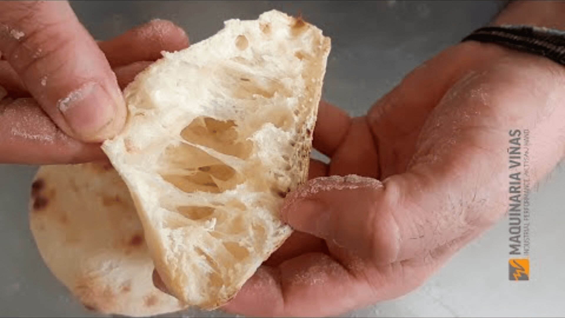 Eine Nahaufnahme von zwei Händen, die ein Stück luftiges, rustikales Brot halten, das seine offene, flauschige Krume und Löcher zeigt. Der Hintergrund ist unscharf, und auf der rechten Seite des Bildes befindet sich das Logo von Maquinaria Viñas.