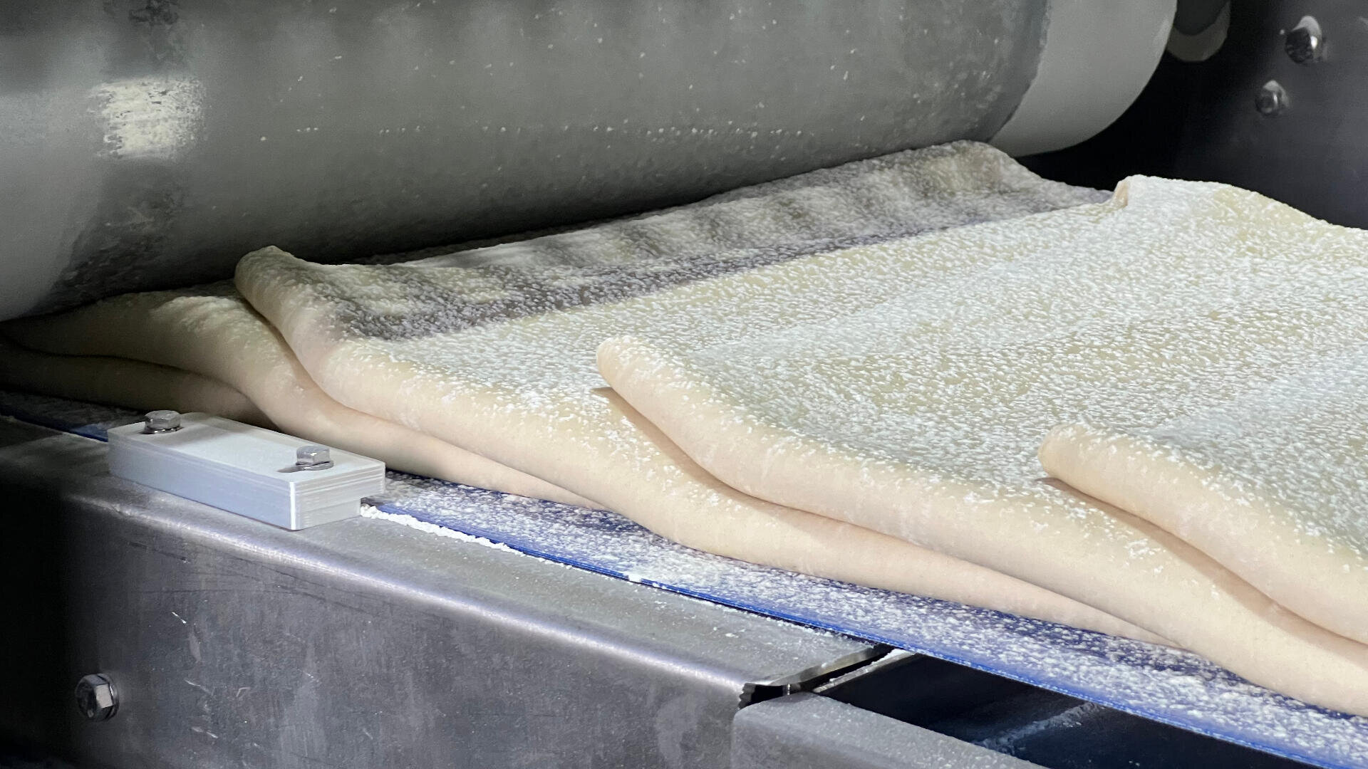 Sheets of dough are being flattened and folded by industrial rollers on a metal conveyor belt in a commercial bakery setting.