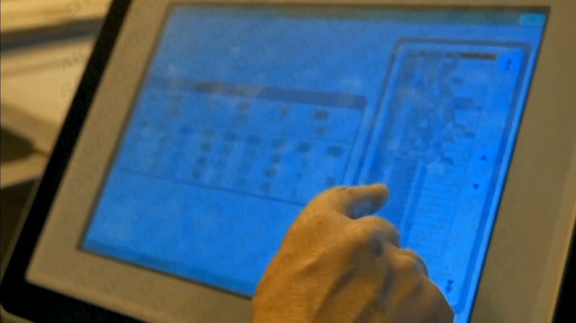 A hand interacts with a touchscreen device displaying a blue interface with various grids, icons, and charts. The screen appears to show data or options in a tabular format.