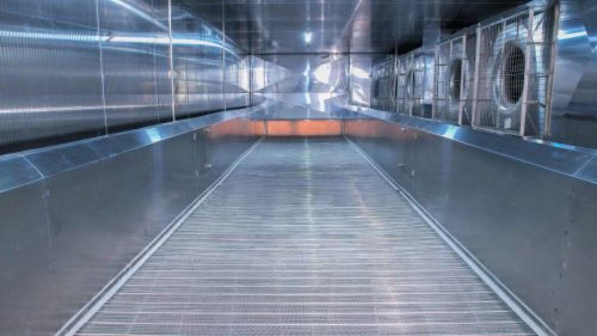 Ein breiter, metallischer, industrieller Flur mit geriffeltem Boden, reflektierenden Wänden, großen Ventilatoren auf der rechten Seite und hellen Lichtern, die eine saubere, futuristische Atmosphäre schaffen.