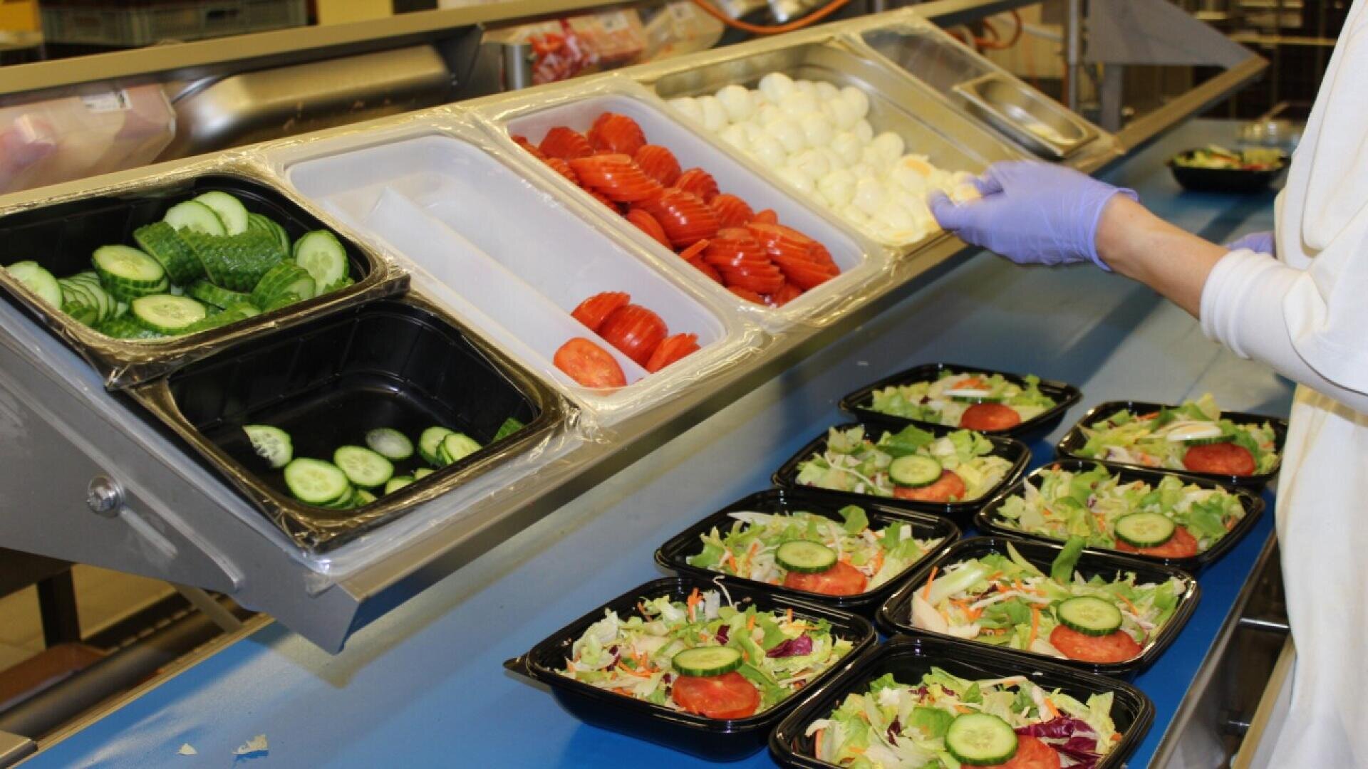 A person wearing gloves prepares salads, placing sliced cucumbers and tomatoes on top. Trays of cucumbers, tomatoes, and hard-boiled eggs are arranged beside prepared salad containers on a counter.