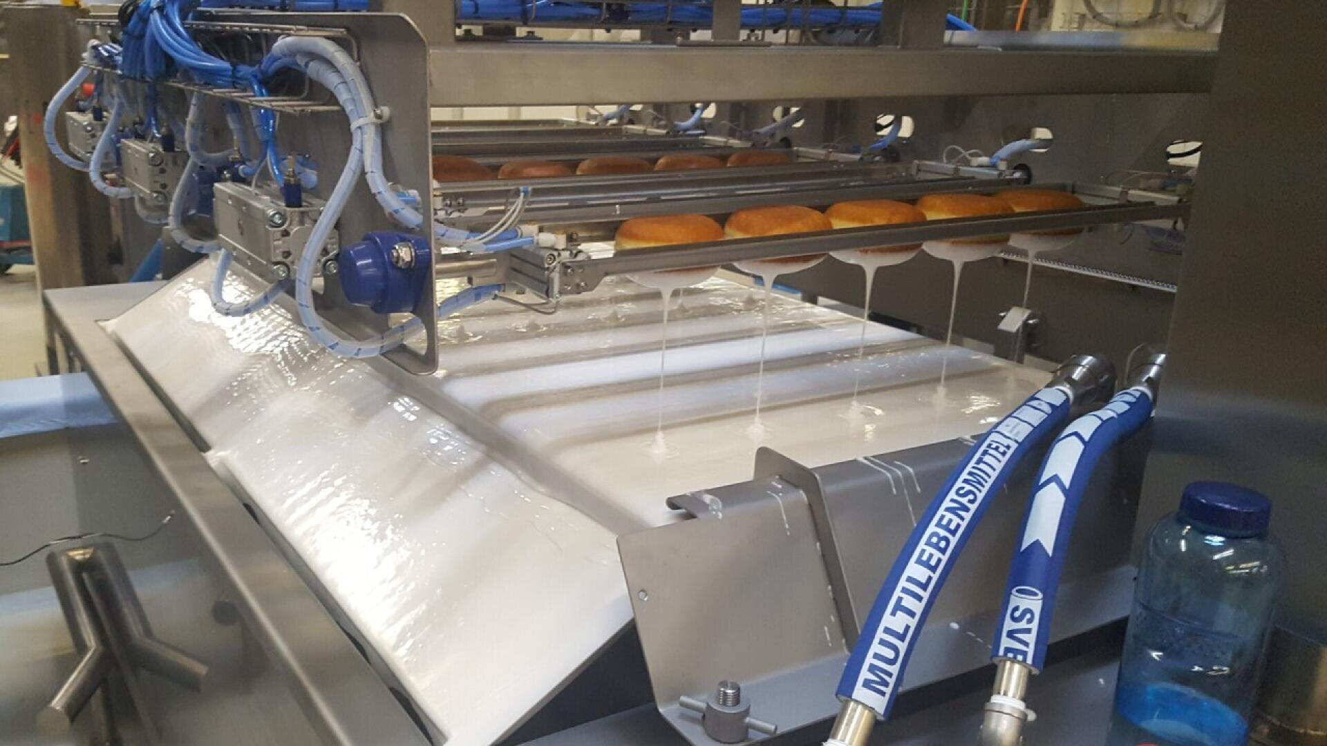 A machine glazes rows of donuts by dripping white icing over them as they move along a conveyor belt in a factory setting. Blue tubes and mechanical parts are visible around the equipment.