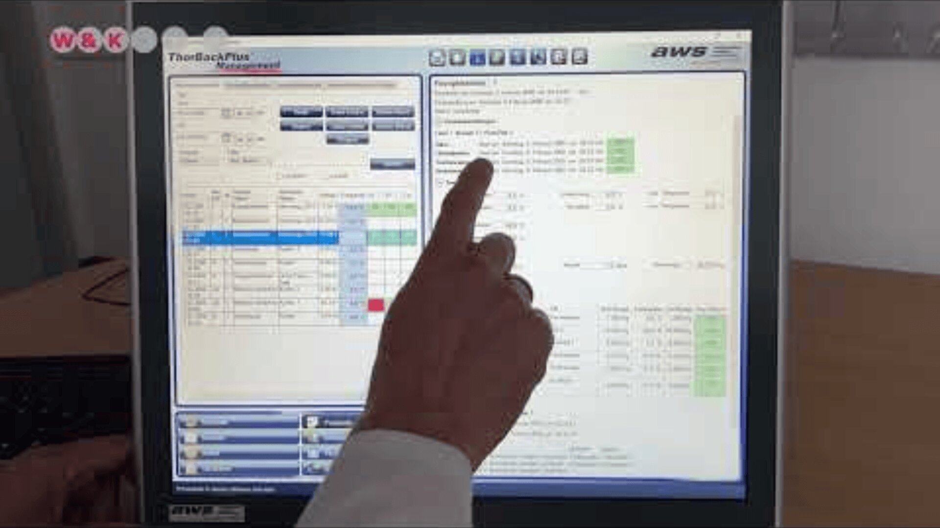 A person’s hand is pointing at a computer monitor displaying a software interface with tables, charts, and highlighted data fields. The screen shows various menus, tabs, and information in a professional setting.