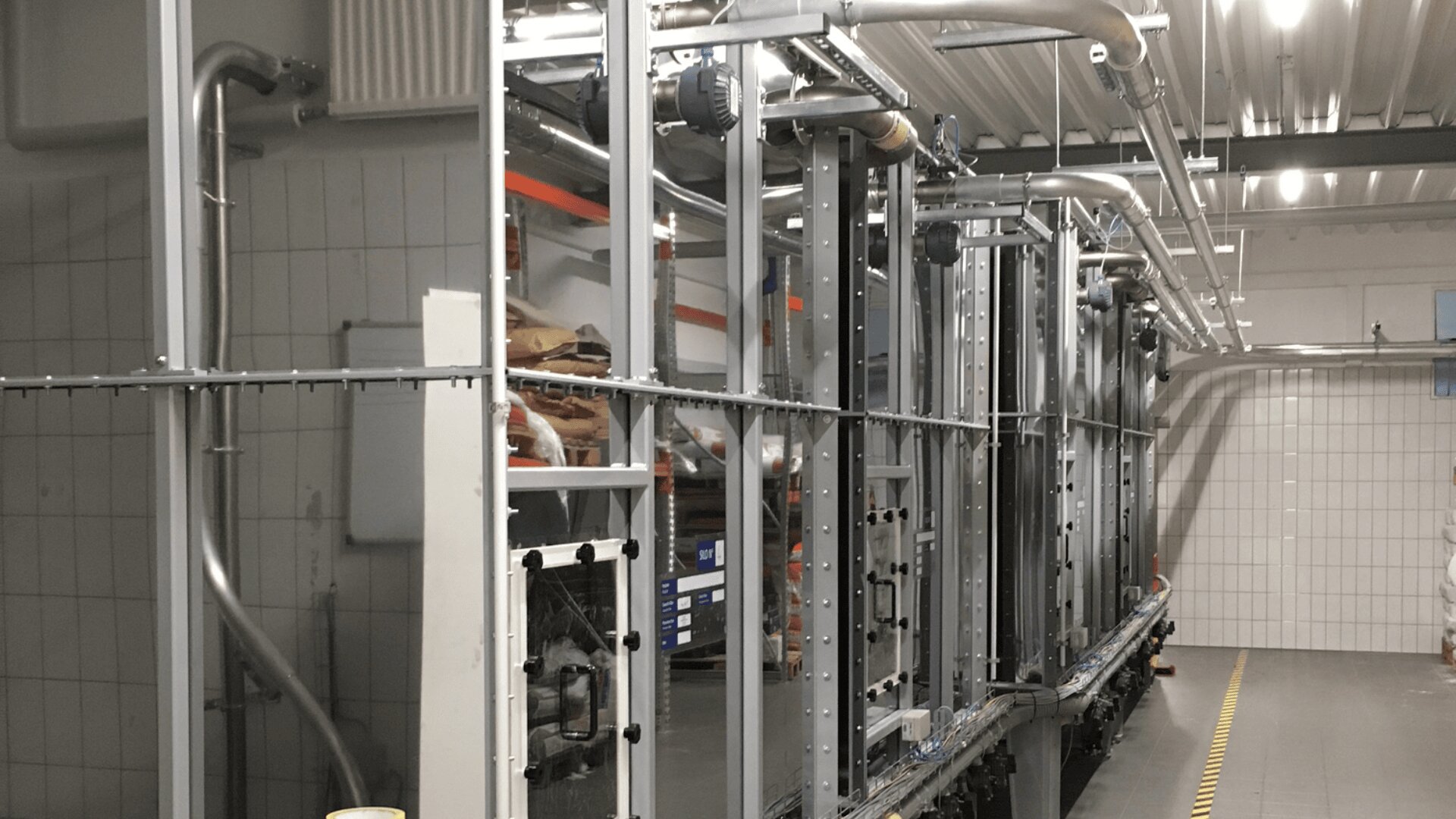 Industrielle Ausrüstung und Metallgerüst in einer gut beleuchteten Fabrik oder Produktionsstätte, mit Rohren, Maschinen und gekachelten Wänden auf der linken Seite.