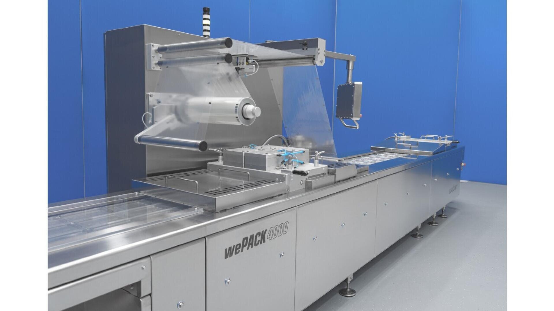 A stainless steel industrial packaging machine labeled wePACK 4000 stands in a clean, blue-walled room, with clear plastic film unspooled and mechanisms for sealing and conveying products.