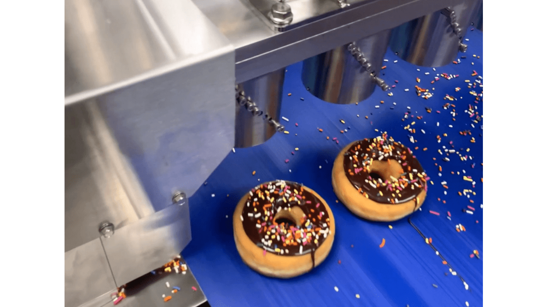 Zwei Donuts mit Schokoladenglasur und bunten Streuseln bewegen sich auf einem blauen Förderband unter einer Edelstahlmaschine, die weitere Streusel ausgibt.