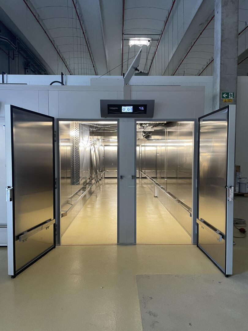 Double doors open to a large, empty industrial cold storage room with stainless steel walls and bright overhead lighting. The floor is yellow, and a digital control panel is above the entrance.
