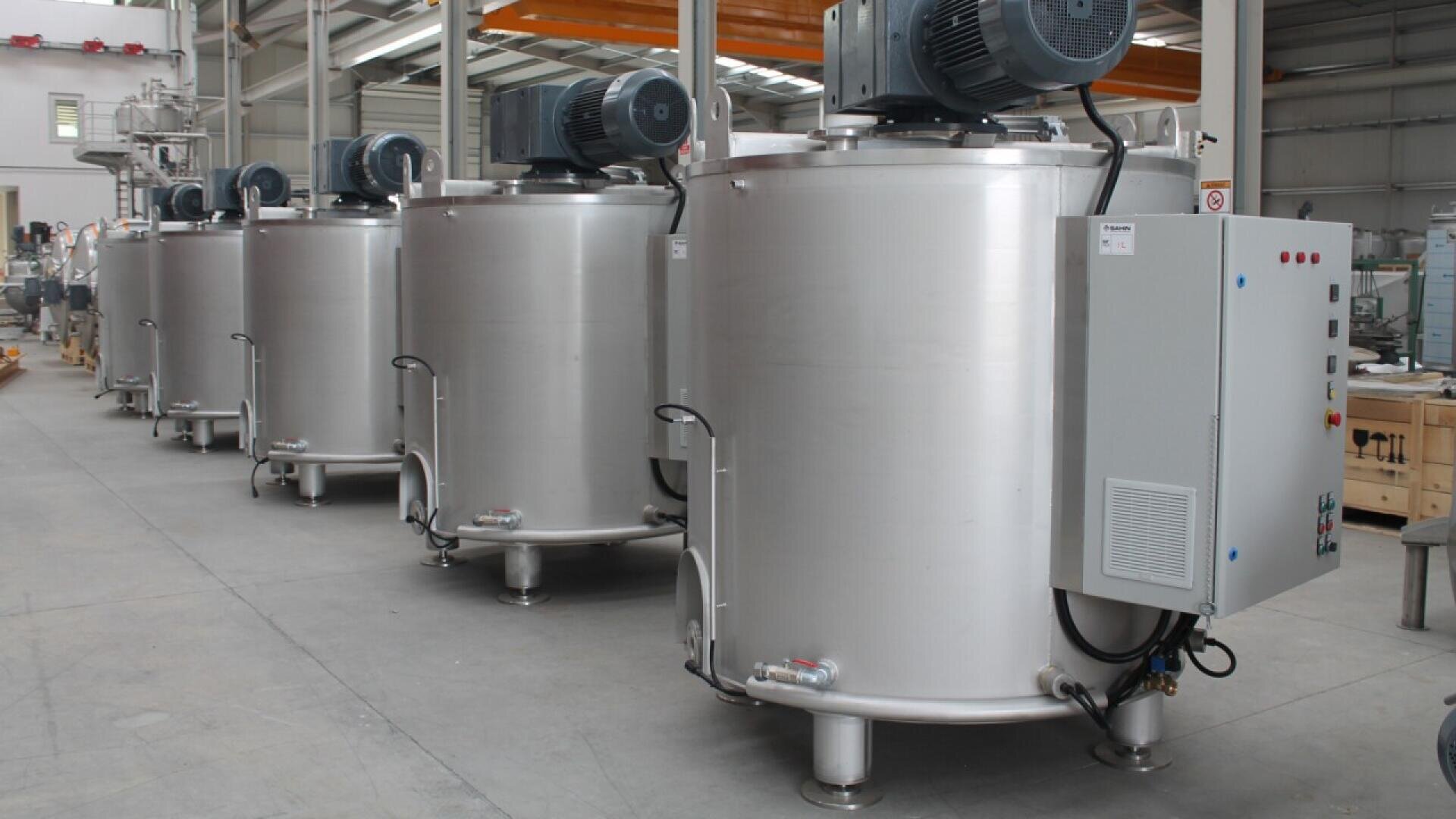 Four large stainless steel industrial mixing tanks with electric motors and control panels, lined up inside a spacious factory with concrete floors and high ceilings.