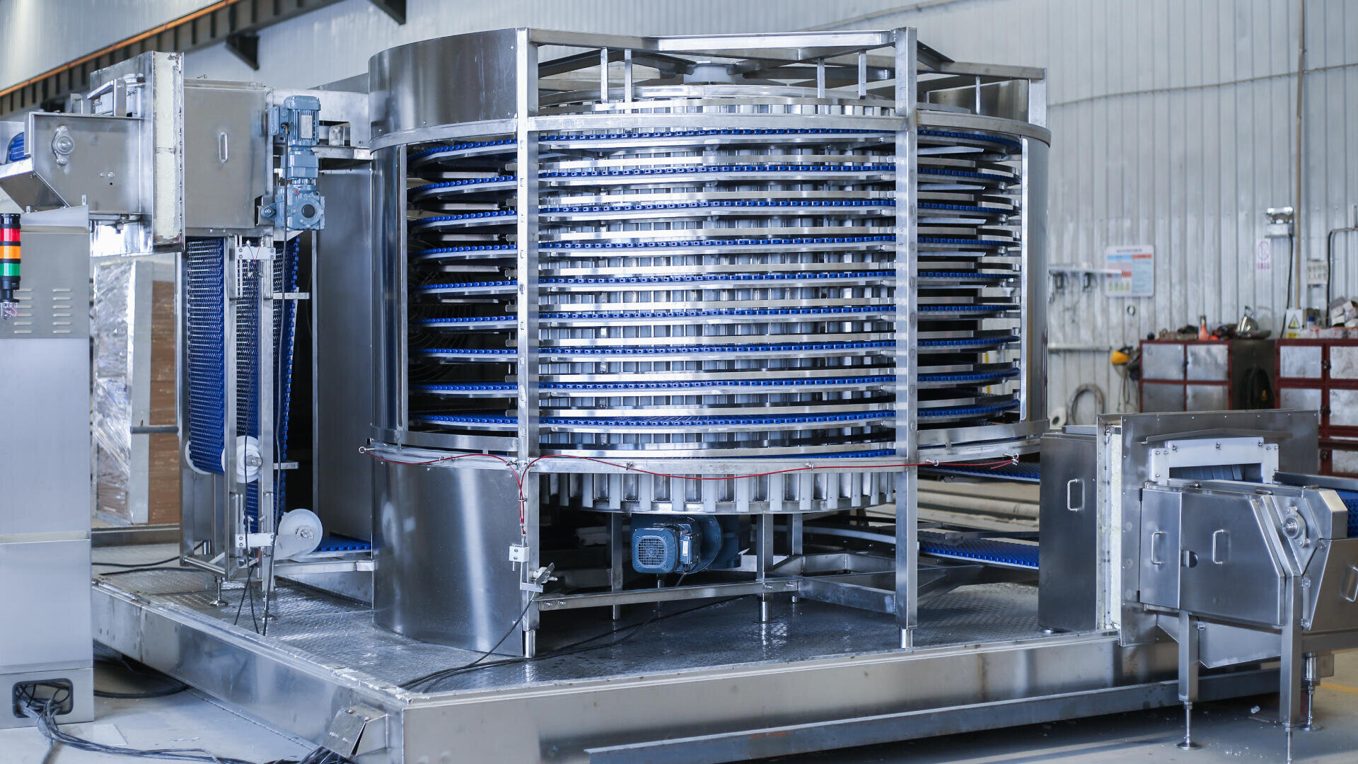 A large industrial spiral conveyor system made of stainless steel sits in a factory setting, with multiple blue belts winding upward around a central column, used for transporting or cooling products.