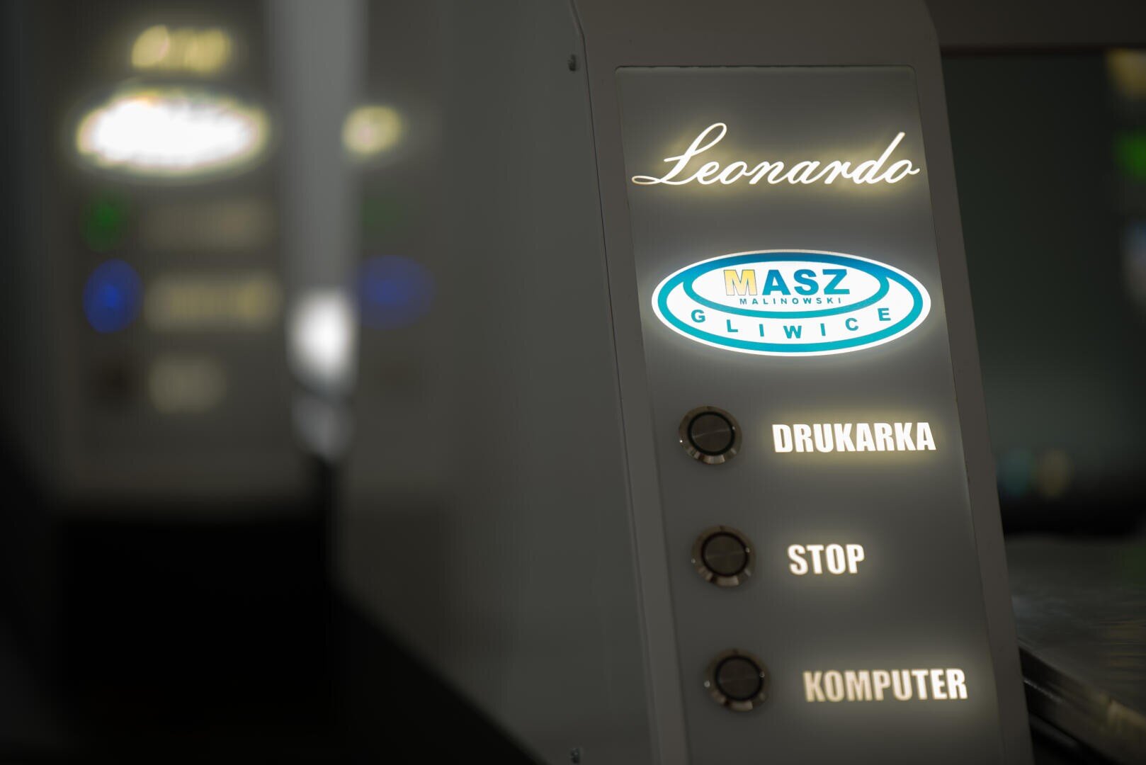 Nahaufnahme eines Bedienfelds mit der Aufschrift „Leonardo“ mit beleuchtetem MASZ Gliwice-Logo und drei Tasten mit den Aufschriften „DRUKARKA“, „STOP“ und „KOMPUTER“. Im Hintergrund ist ein ähnliches Bedienfeld unscharf dargestellt.