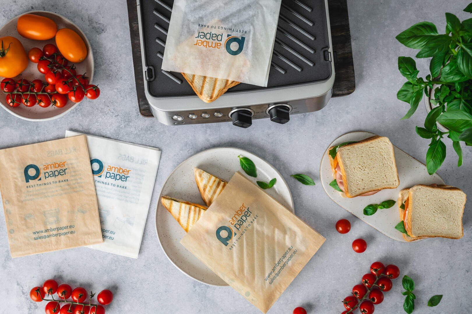 Draufsicht auf gegrillte Sandwiches in bernsteinfarbenen Papierhüllen neben frischen Tomaten, Blattgemüse, einer Panini-Presse und Sandwich-Zutaten auf einer grauen Arbeitsplatte.