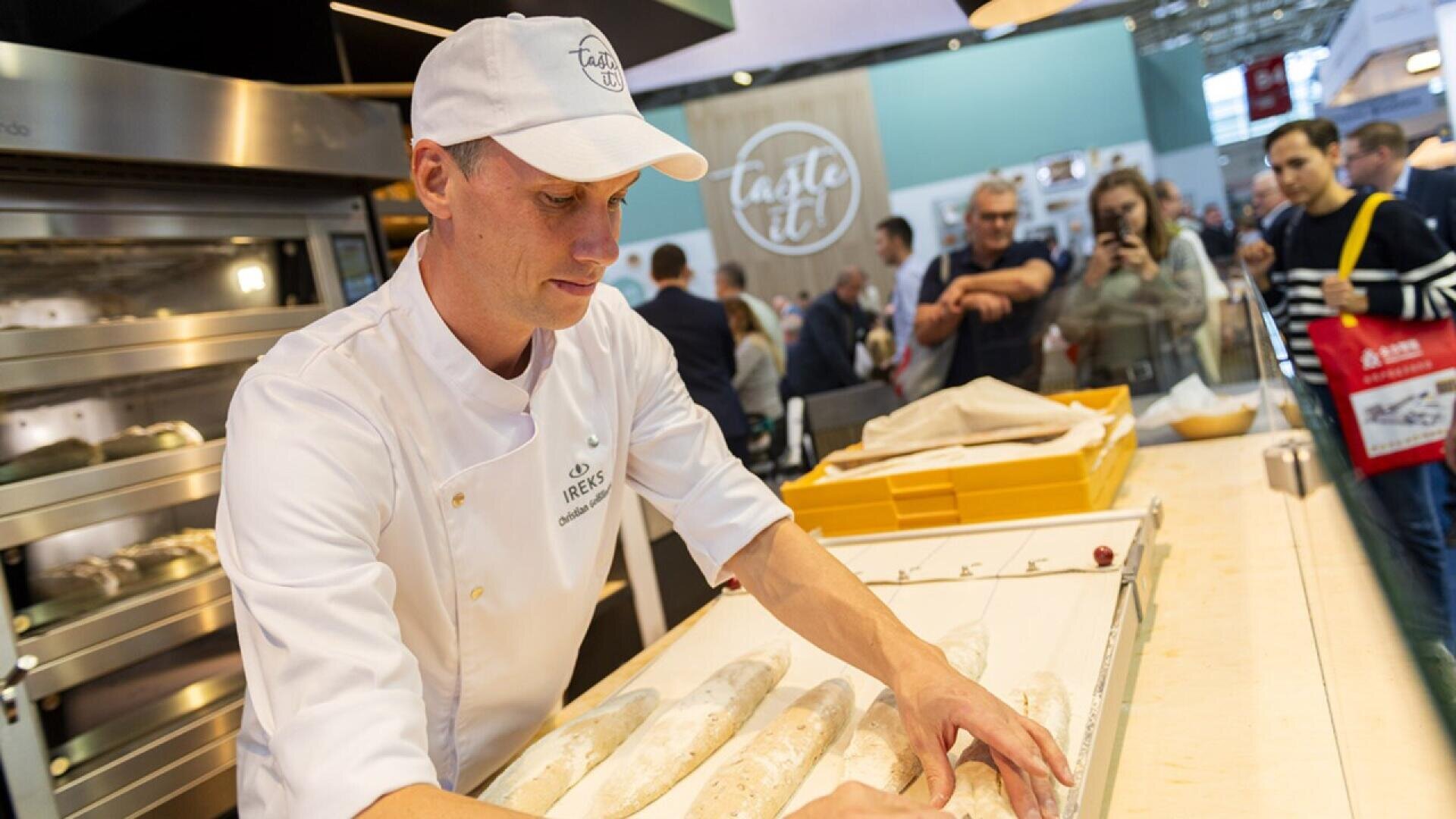 A baker in a white uniform and cap shapes baguette dough on a counter at a public event, with several people watching and taking photos in the background. Baked goods and ovens are visible nearby.