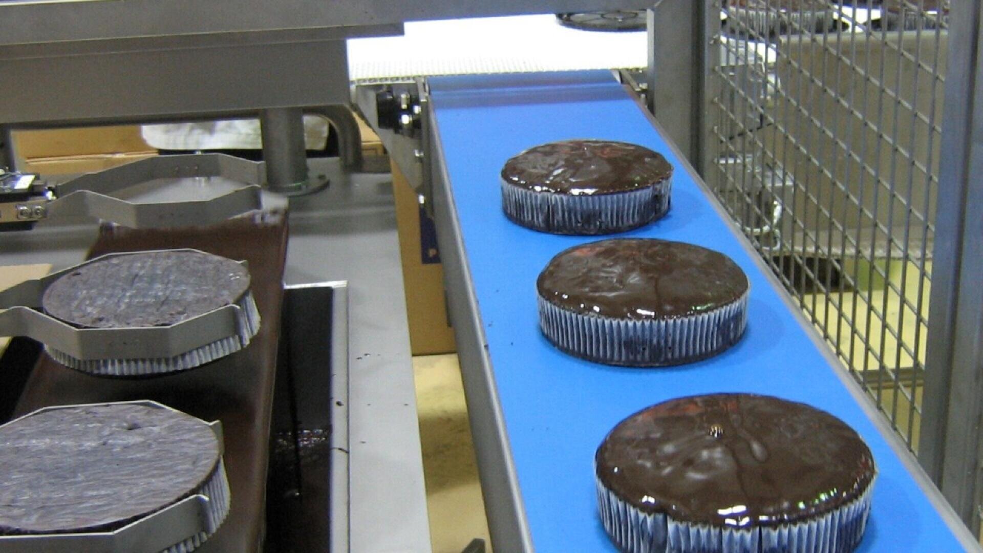 Chocolate cakes covered in shiny chocolate glaze move along a blue conveyor belt in a factory setting, with metal machinery visible in the background.