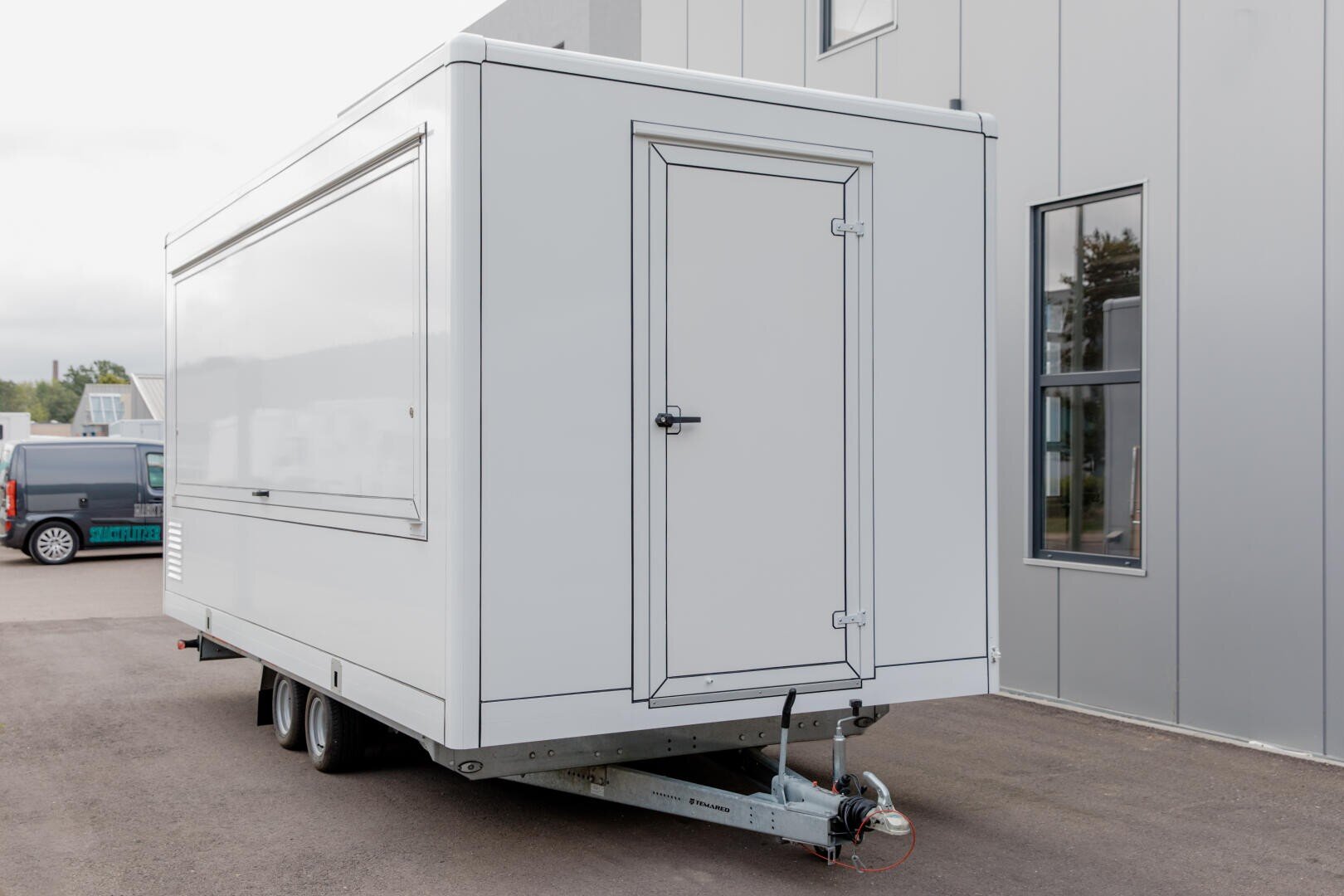 Ein weißer mobiler Anhänger mit einer geschlossenen Tür und einem Fenster, der auf einer gepflasterten Fläche neben einem modernen Gebäude mit großen Fenstern steht. Der Anhänger steht auf Rädern und hat an der Vorderseite eine Anhängerkupplung zum Ziehen.