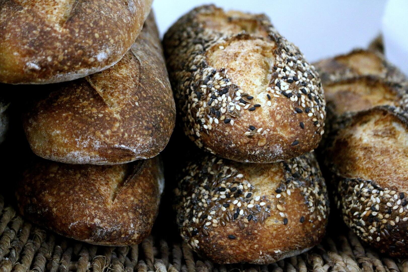 A close-up of rustic bread loaves, some topped with a mix of seeds, stacked on a woven mat. The bread has a golden-brown crust and a slightly cracked surface.
