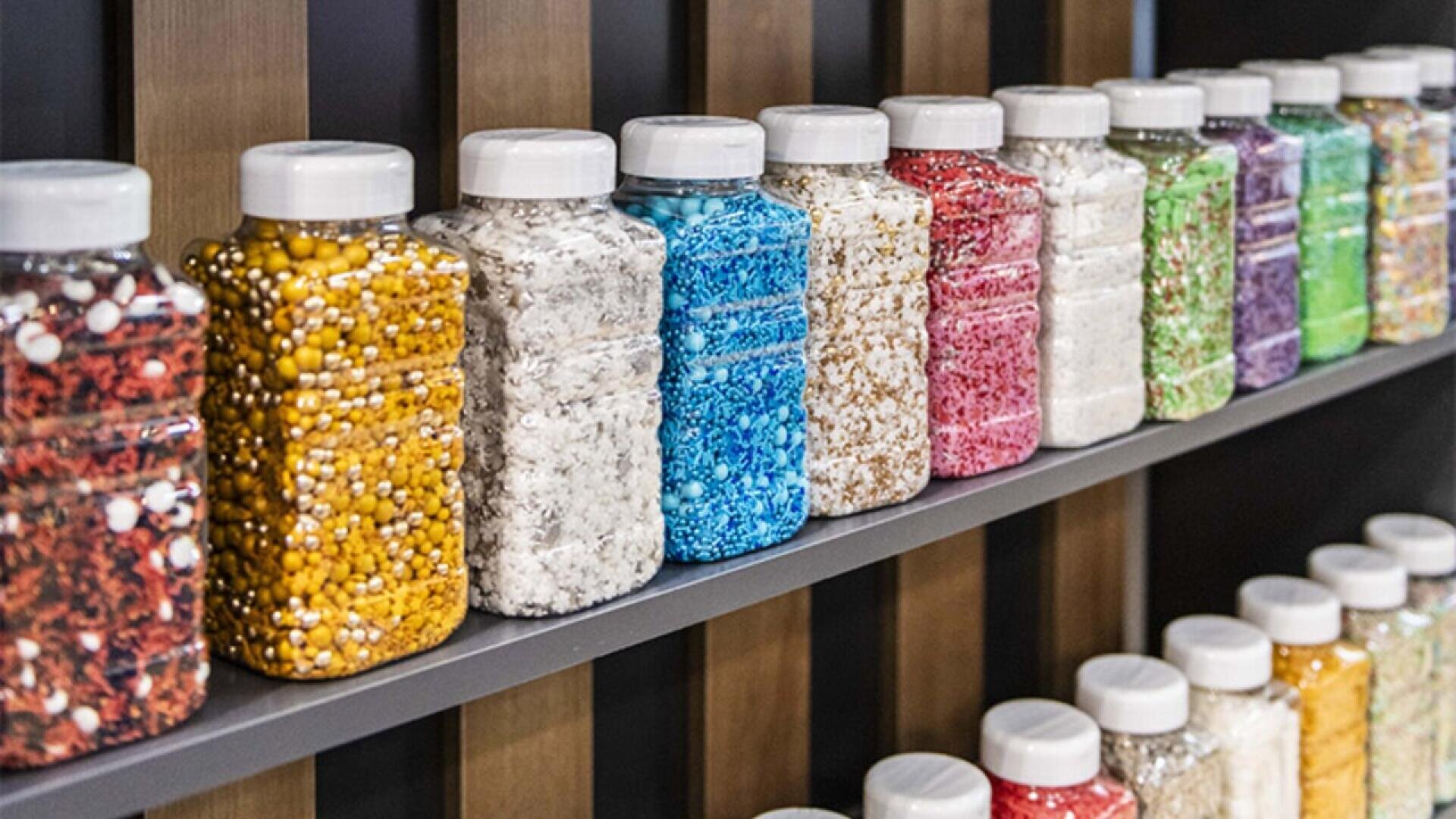 Plastic jars filled with various colorful sprinkles and baking decorations are lined up on two shelves, arranged in a spectrum from red and yellow to green and purple, with white lids on each jar.