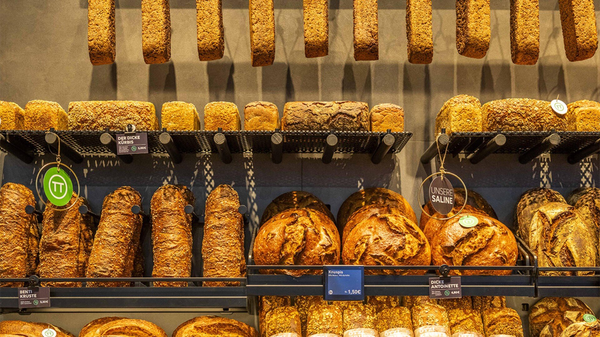 In einer Bäckerei sind verschiedene Arten von handwerklich hergestellten Broten in Regalen an der Wand ausgestellt, mit Preisschildern und Etiketten, die die verschiedenen Sorten und Zutaten angeben.