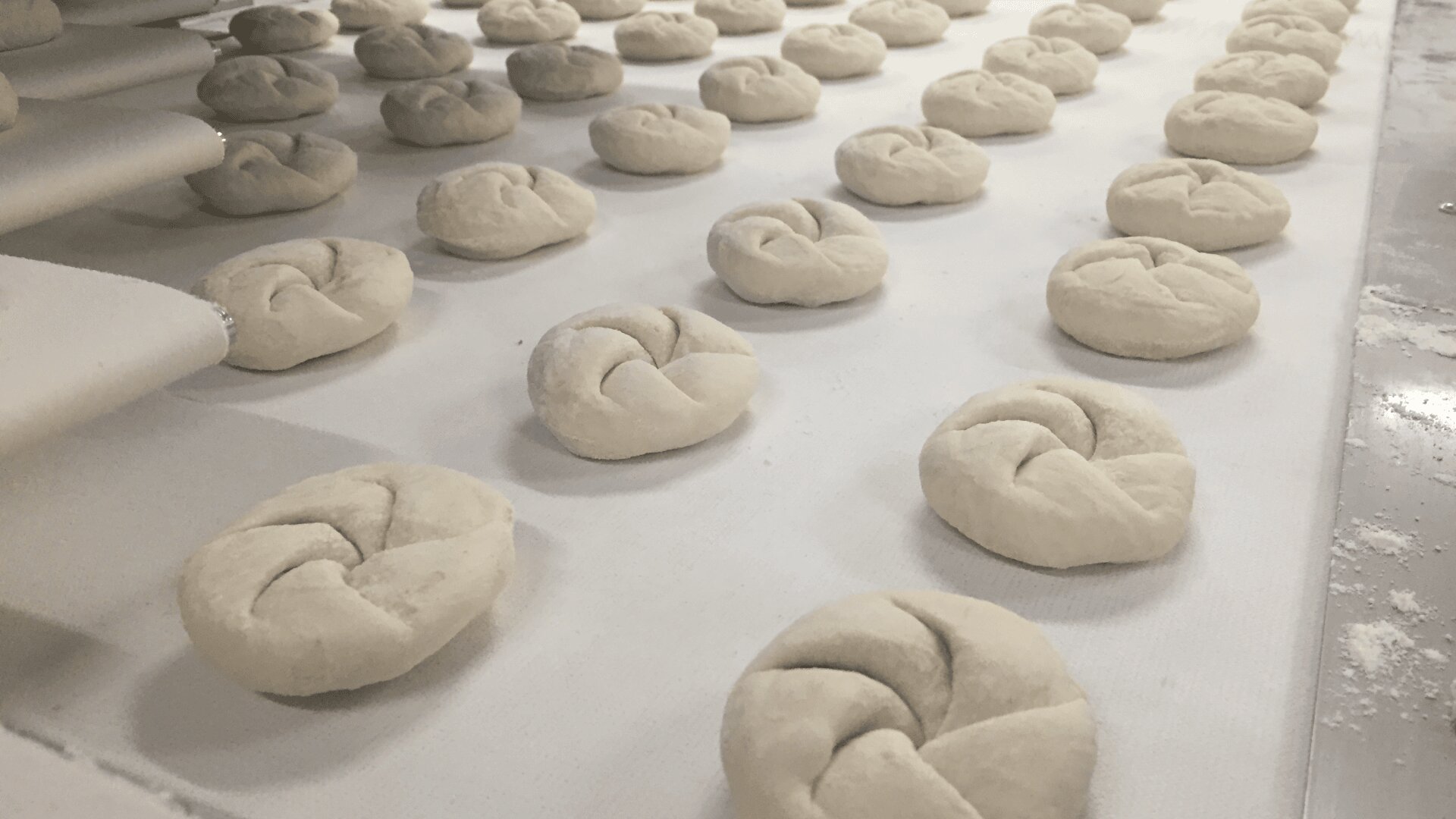 Dozens of unbaked, knotted dough rolls are arranged in neat rows on a white surface, ready for baking. The background includes a conveyor system, suggesting a commercial or industrial bakery setting.