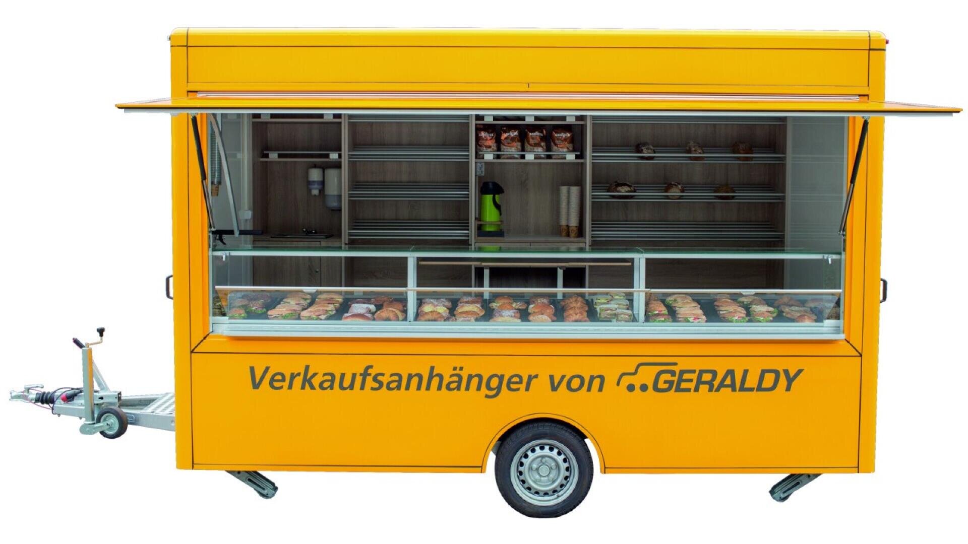 A yellow food trailer with large display windows showing pastries and bread. The trailer reads Verkaufsanhänger von GERALDY and has shelves with more baked goods inside.
