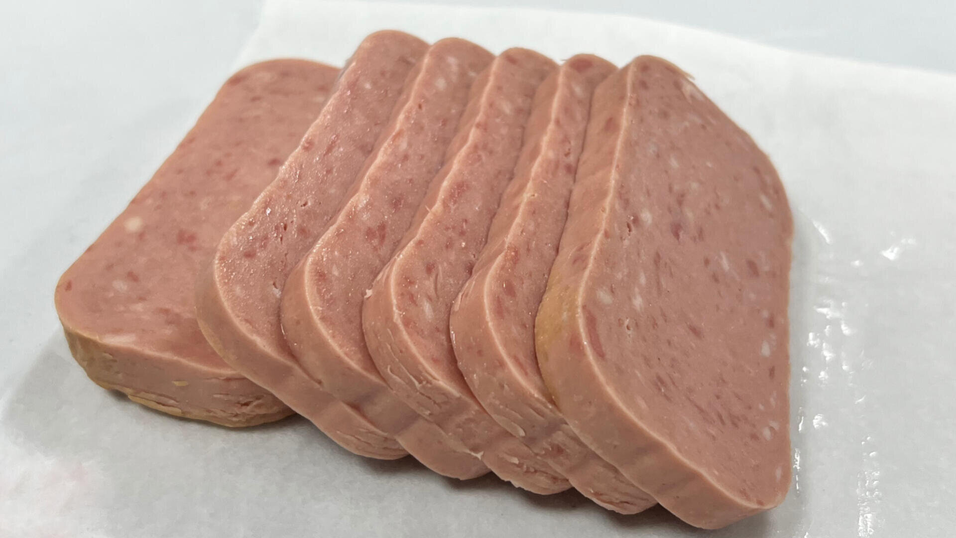 Six slices of processed luncheon meat, such as Spam, are neatly arranged on a sheet of white parchment paper against a plain background.
