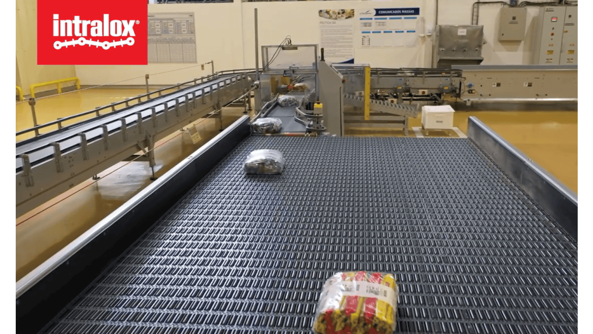 Ein Förderband in einer Fabrik transportiert verpackte Lebensmittel. Im Hintergrund sind Maschinen und Schalttafeln zu sehen. Das intralox-Logo erscheint in der oberen linken Ecke.