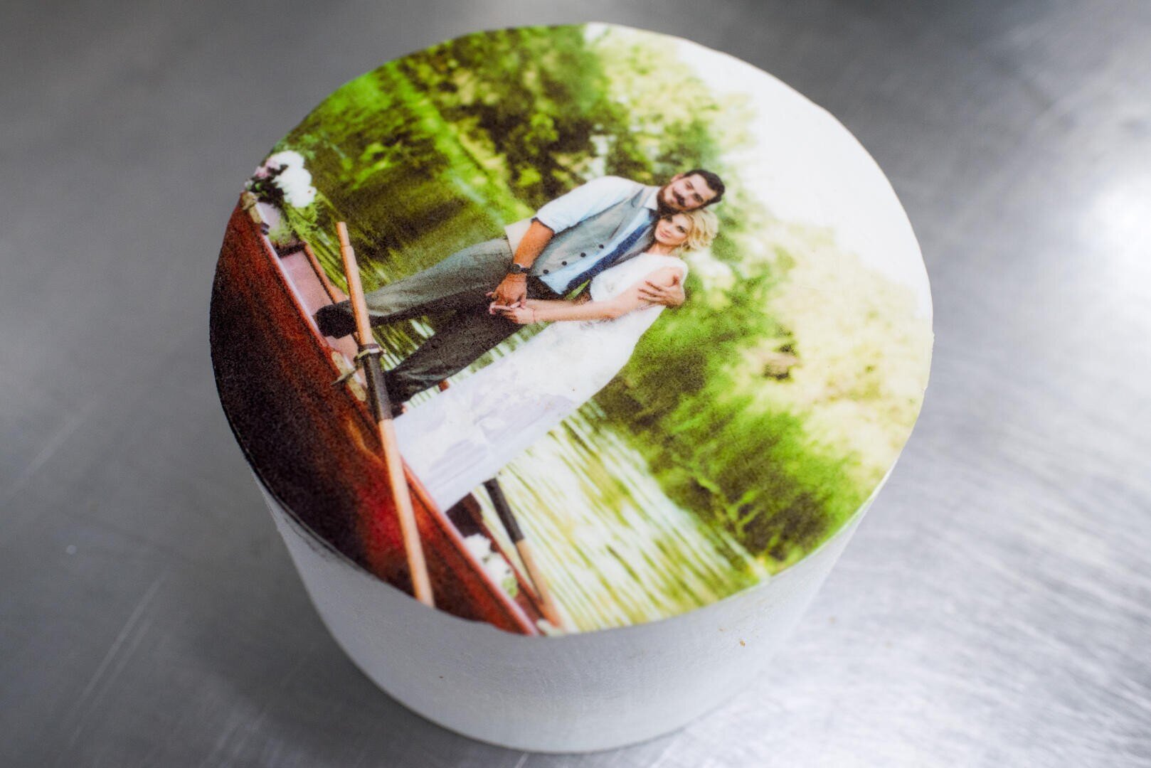 Ein Kuchen, gekrönt mit einem essbaren gedruckten Foto eines lächelnden Paares in Hochzeitskleidung, das auf einem Holzsteg an einem grünen, von Bäumen gesäumten See steht.