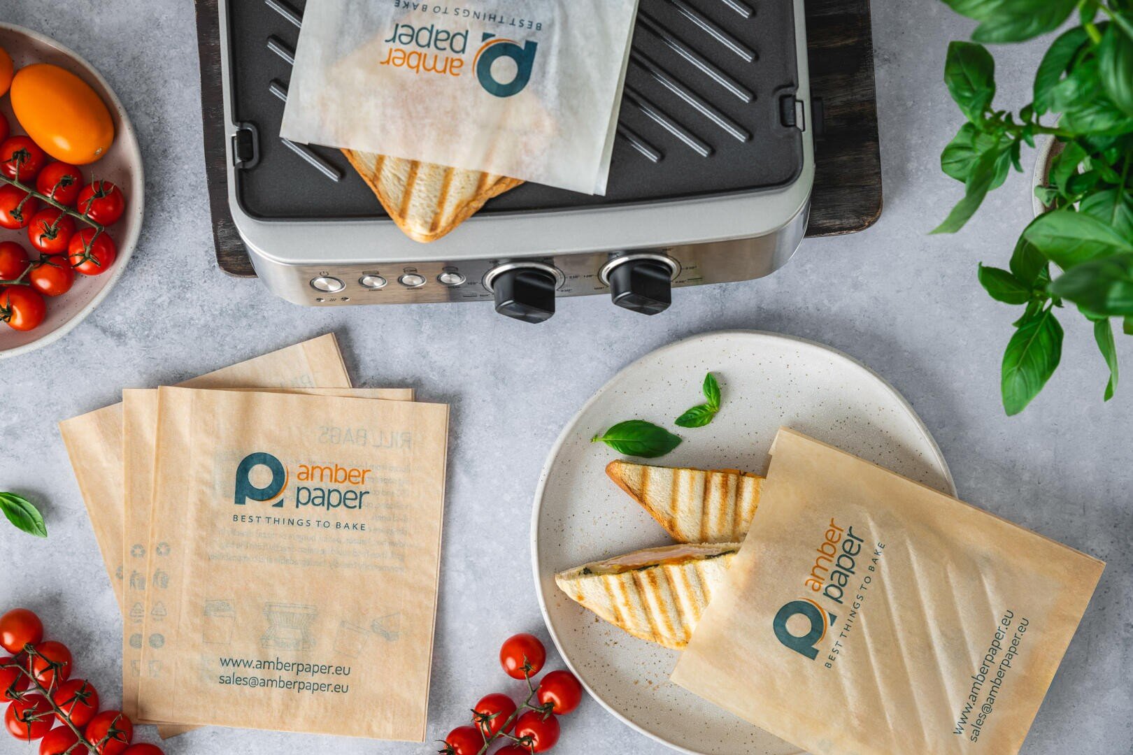 A plate with two grilled sandwiches in crisp paper sleeves sits beside a grill, fresh cherry tomatoes, basil, and more sandwich sleeves on a table, creating a fresh and inviting meal setting.