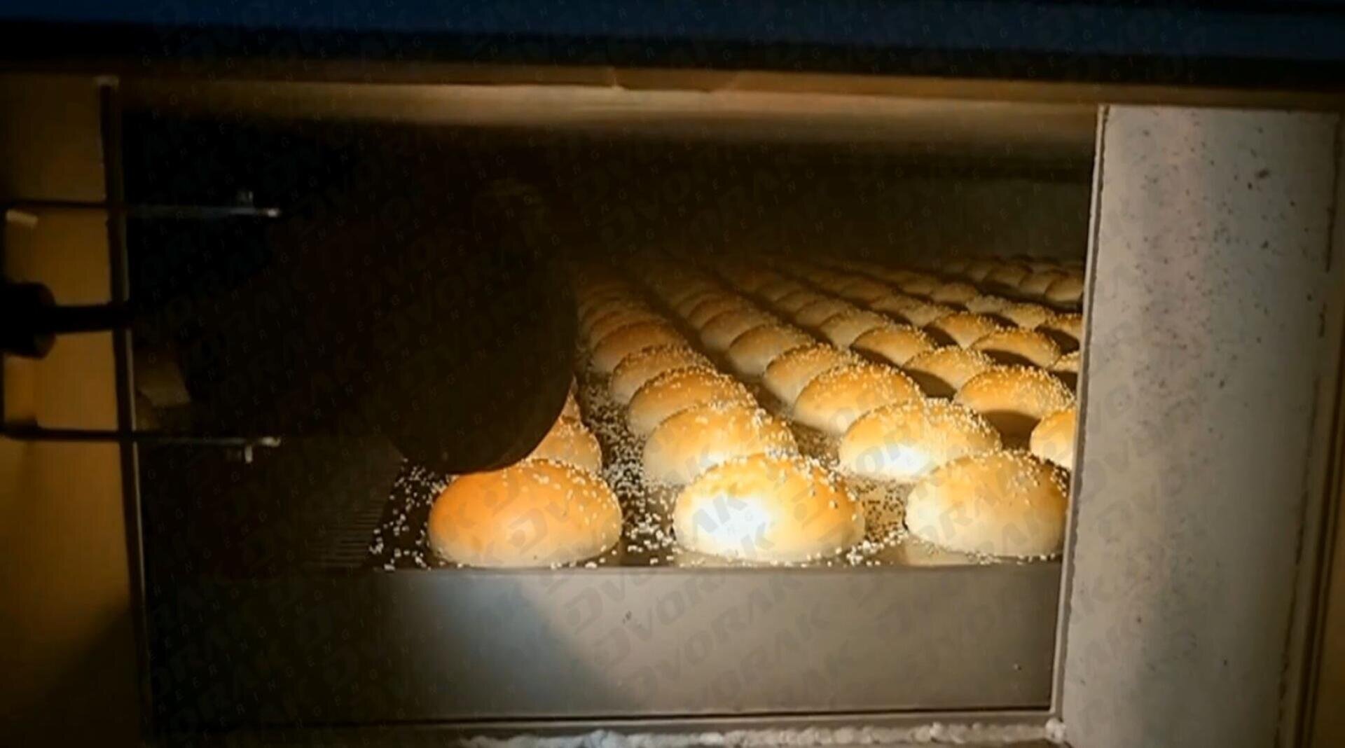 Rows of golden brown bread rolls sprinkled with sesame seeds are baking inside an industrial oven, illuminated by the oven light.