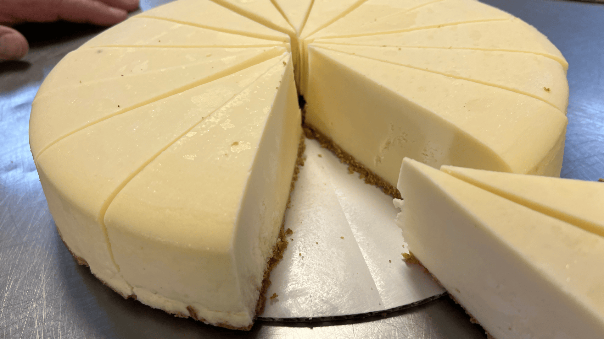 A round, sliced cheesecake with a smooth, creamy top and a graham cracker crust sits on a metal surface. One slice is being removed, revealing the cakes dense, rich texture.