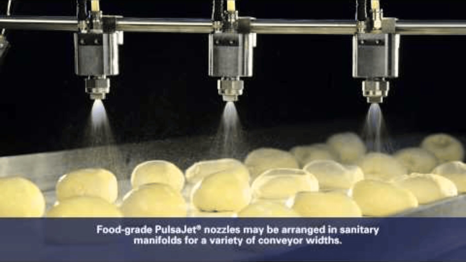 Reihen ungebackener Teigbällchen auf einem Förderband werden von mehreren lebensmittelechten PulsaJet®-Düsen besprüht, die in einer hygienischen Produktionsumgebung Nebel verteilen. Eine Bildunterschrift beschreibt die flexible Anordnung der Düsen.
