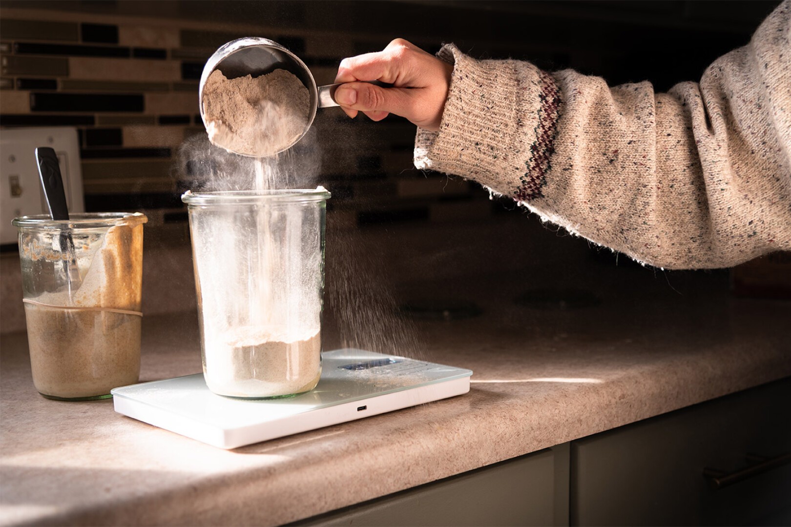 Eine Person schüttet Mehl aus einem Metallmessbecher in ein Glasgefäß, das auf einer digitalen Küchenwaage auf einer Arbeitsplatte steht. Ein weiteres Glas, teilweise mit Teig bedeckt, steht daneben. Sonnenlicht fällt auf die Szene.