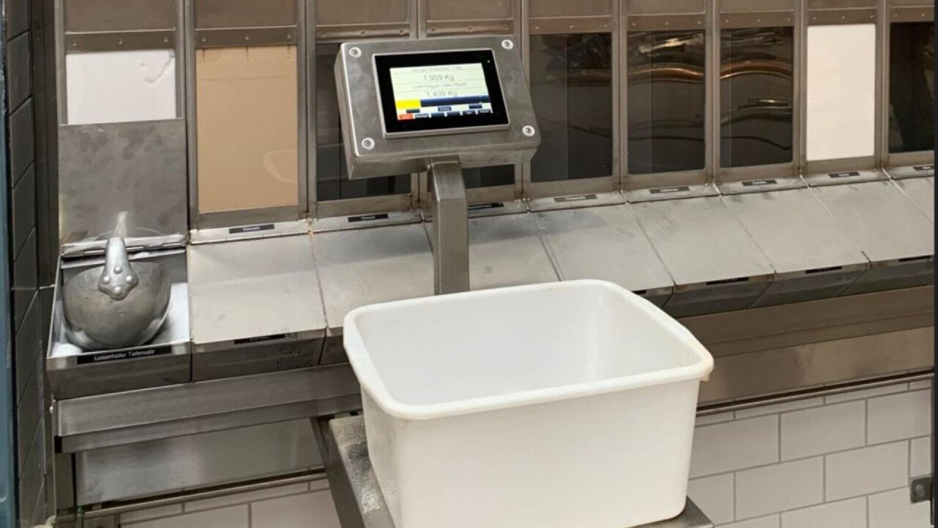 A digital scale with a display screen is mounted above a stainless steel counter, holding an empty white plastic bin. A metal container with a bird-shaped handle sits to the left of the scale.