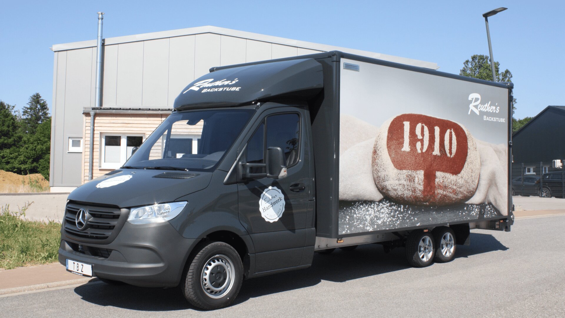 Ein schwarzer Lieferwagen mit dem Schriftzug Raders Backstube und einem Bild von Brot mit 1910 auf der Seite, geparkt auf einer Straße in der Nähe von Industriegebäuden.