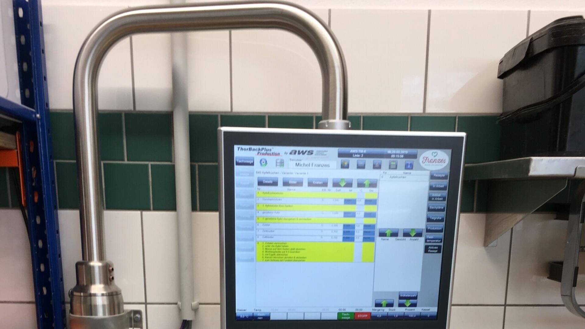 A touchscreen control panel with a metallic frame displays a software interface featuring lists and buttons in yellow, blue, and green, mounted in a tiled industrial workspace with shelves nearby.