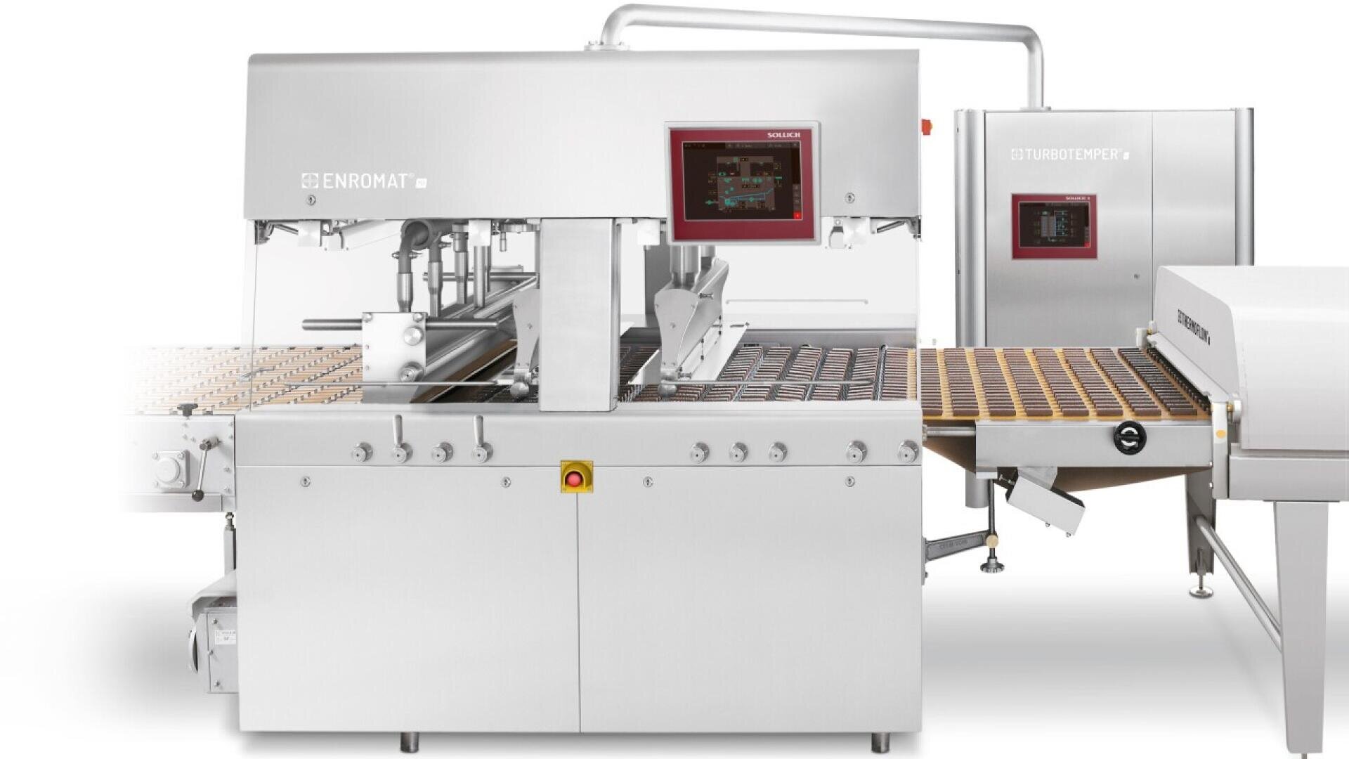 A stainless steel industrial chocolate enrobing machine with conveyor belts and control panels, used for coating confections in chocolate. Brown treats are visible on the belt.