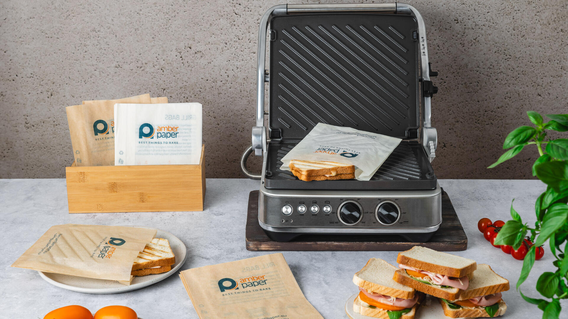 A sandwich press with a grilled sandwich inside sits on a counter. Nearby are sandwiches in brown paper sleeves, a wooden holder with more sleeves, sliced tomatoes, and fresh basil, creating a clean, inviting kitchen scene.