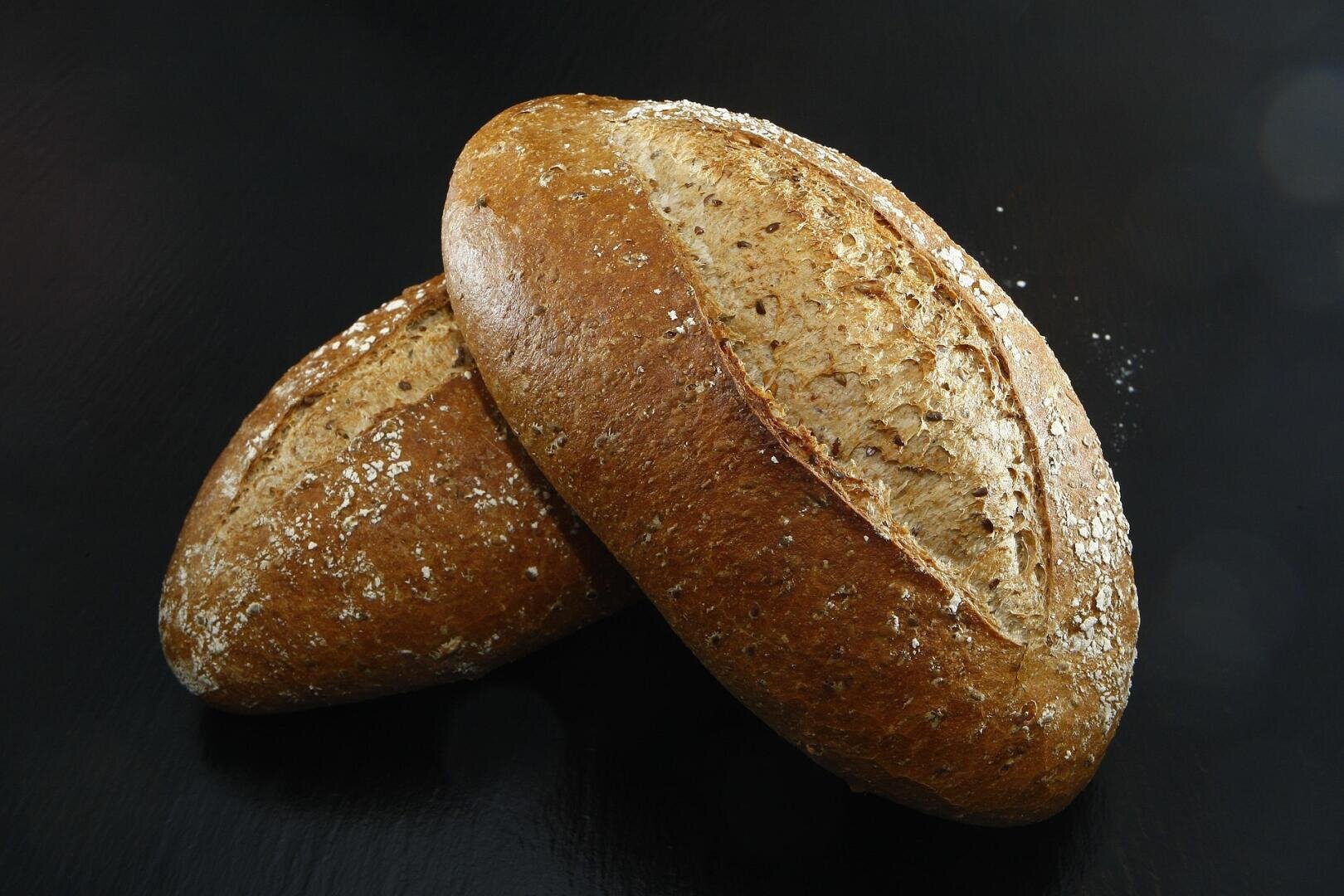 Two oval-shaped loaves of whole grain bread with a golden-brown crust and visible oats rest on a dark, smooth surface.