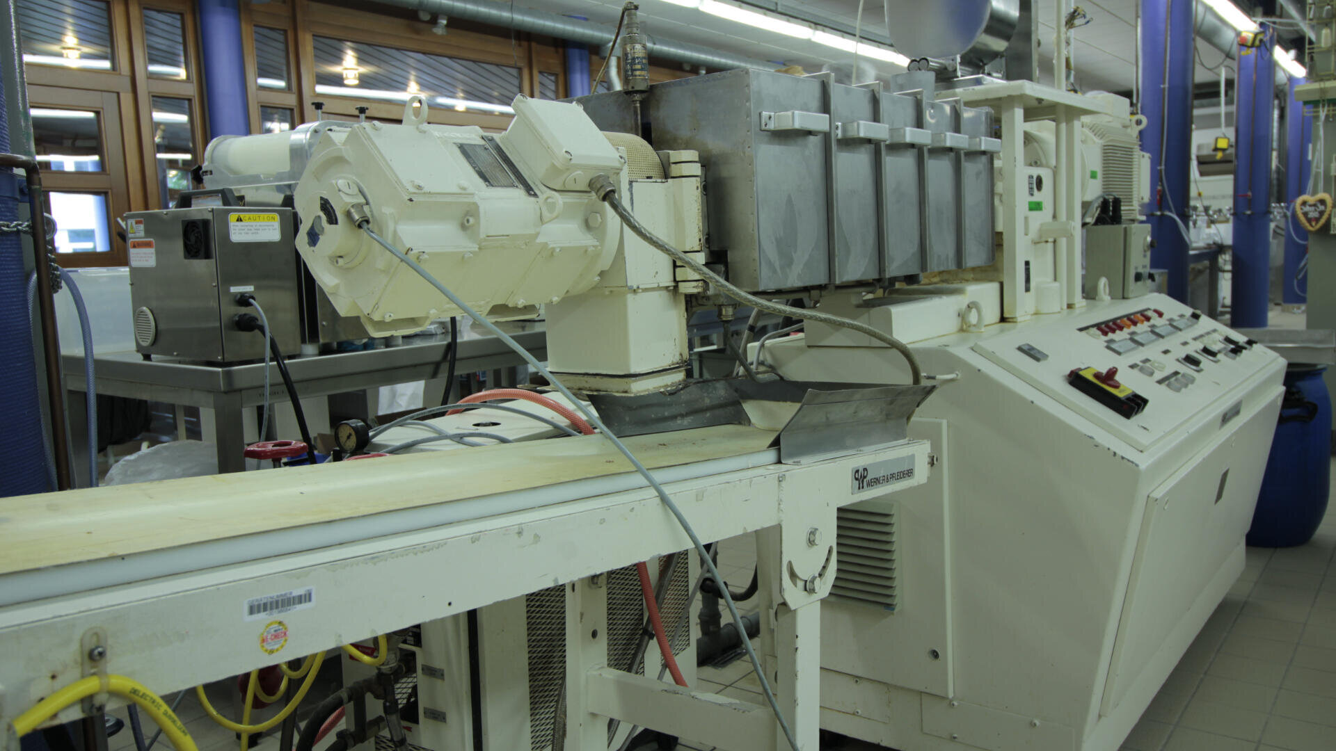Eine große industrielle Extrudermaschine mit Metalltrichtern und Bedienfeldern ist in einer Fabrikumgebung aufgestellt, umgeben von Kabeln und anderen Geräten.